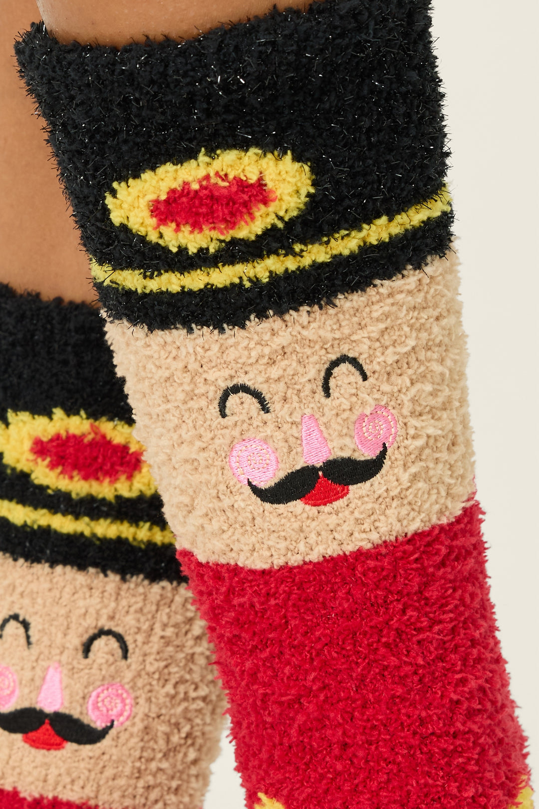 Close-up of P.J. Salvage Ski Jammie Classics Nutcracker Crew Socks with Grippers, featuring a fuzzy design with a smiling nutcracker face, black hat, pink cheeks, mustache, and red and yellow details.