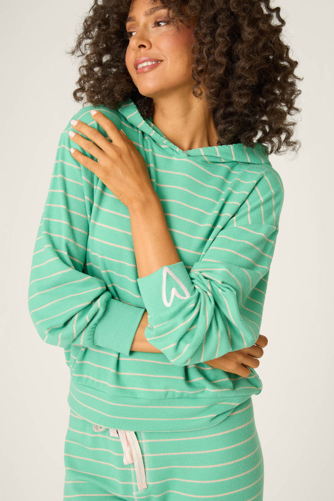 A woman with curly hair wears the P.J. Salvage Peace & Pawlidays Green Stripe Hoodie, featuring festive stripes and a heart sleeve detail. She stands arms crossed, smiling slightly—a cozy winter look.