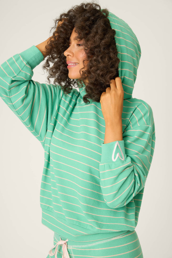 A person with curly hair smiles to the side, wearing the P.J. Salvage Peace & Pawlidays Green Stripe Hoodie with holiday stripes, hood up, and a small heart near the left wrist—perfect for winter lounging.