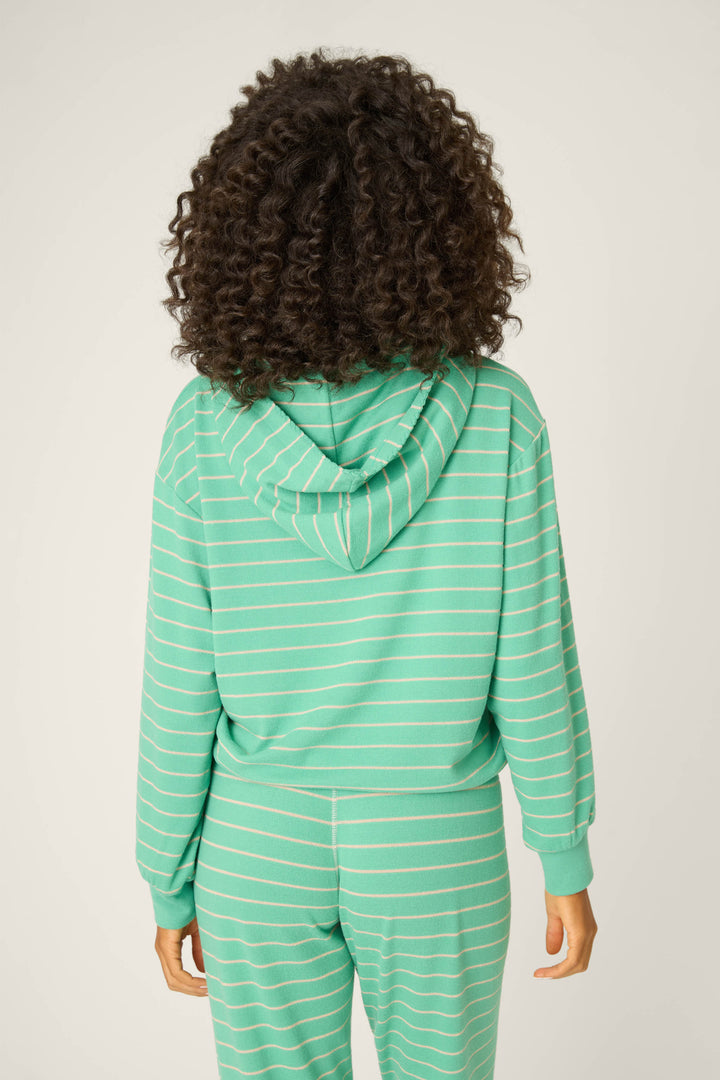 A person with curly hair is shown from the back, wearing the P.J. Salvage Peace & Pawlidays Green Stripe Hoodie with matching pants. The hood is up, ideal for winter lounging, and both hands are relaxed at their sides.