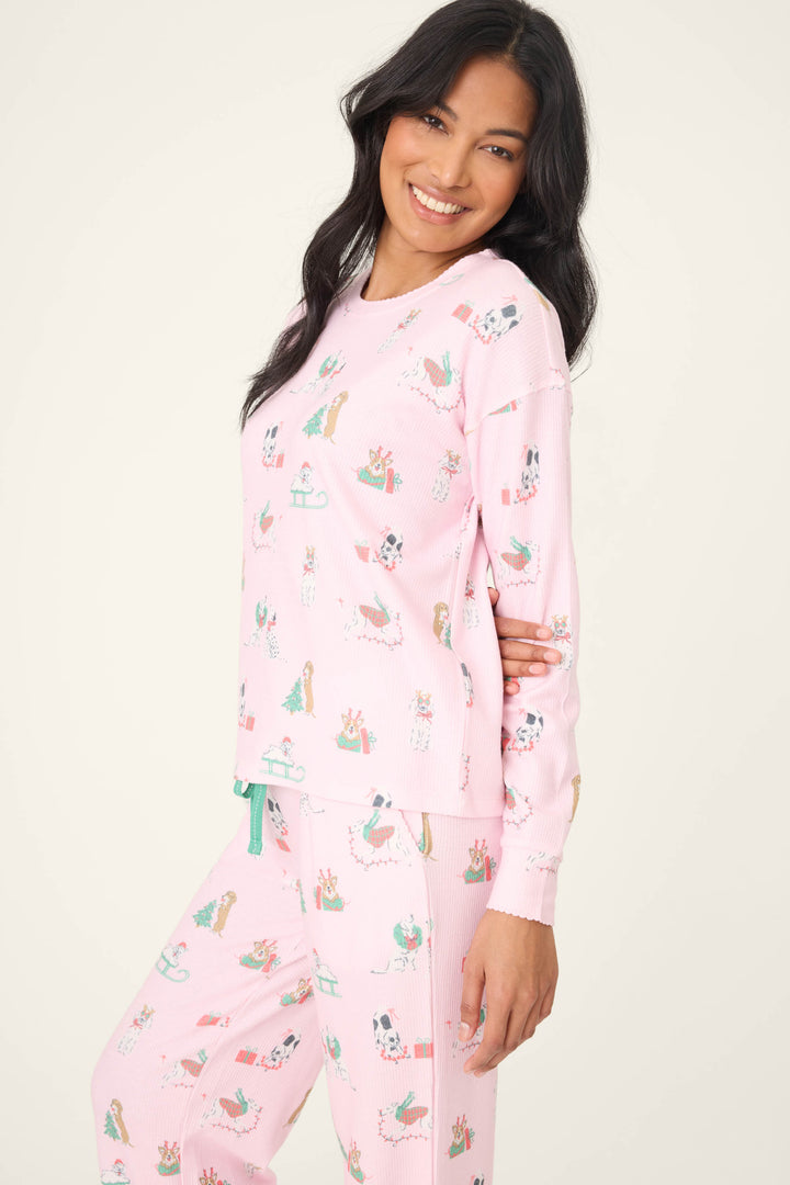A woman with long dark hair smiles at the camera in a P.J. Salvage Peace & Pawlidays Dog Print Long Sleeve Top, featuring playful dog illustrations, standing against a plain light background.