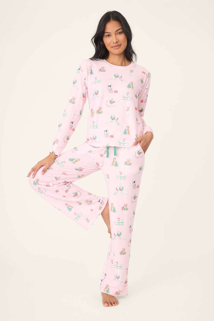 A person stands barefoot on one leg, wearing P.J. Salvage Peace & Pawlidays Dog Print Long Sleeve Top and matching loose pajama pants with pockets and a teal drawstring. The pajamas feature a playful dog print in light pink.