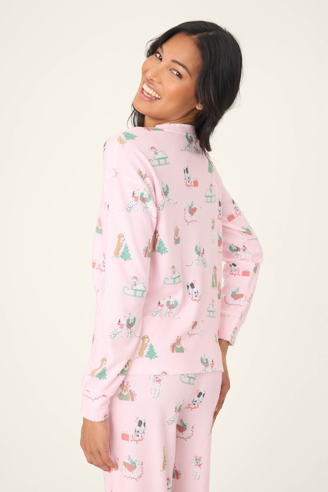 A person with long dark hair smiles over their shoulder, wearing the P.J. Salvage Peace & Pawlidays Dog Print Long Sleeve Top in light pink with dog and holiday motifs, standing against a plain light background.