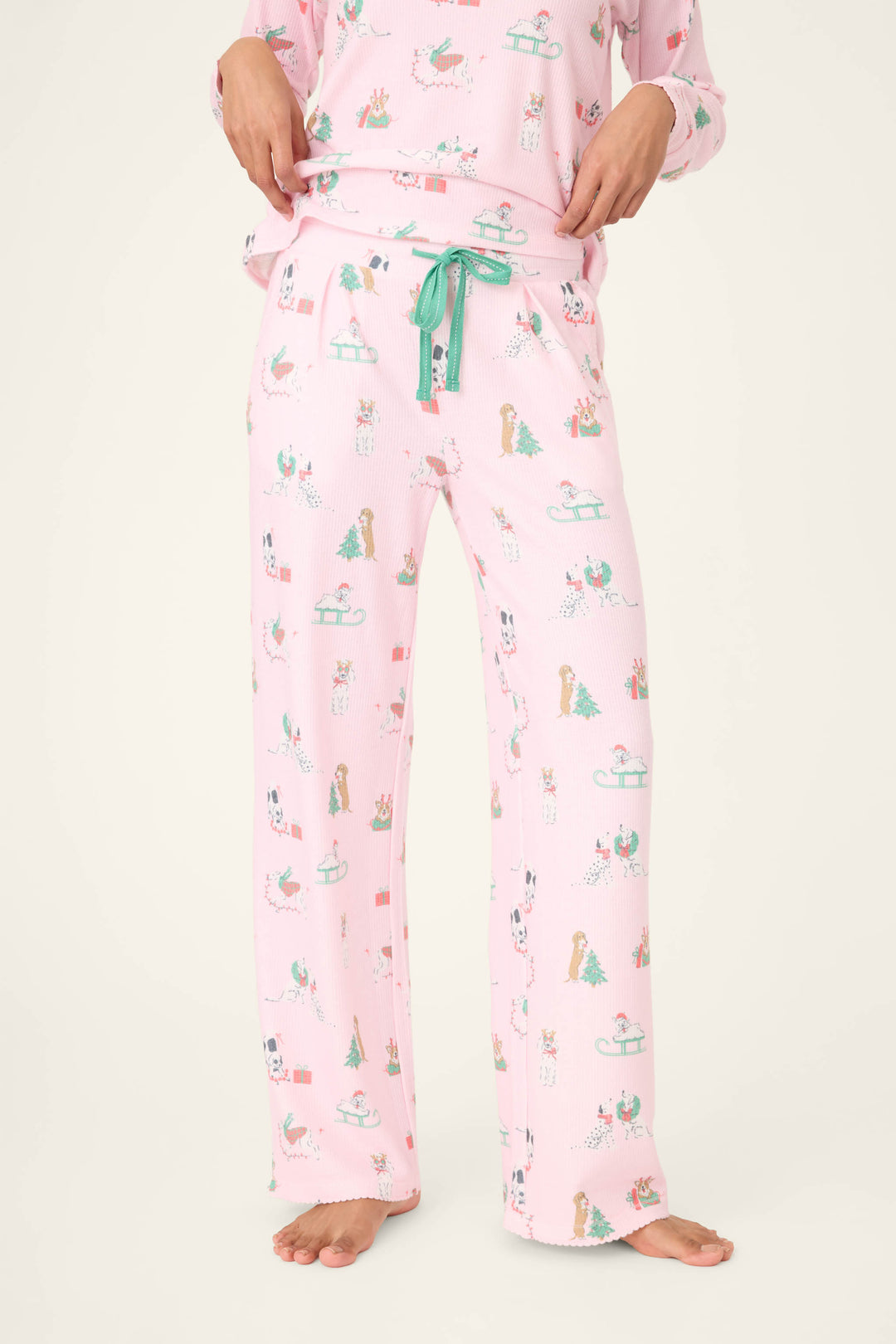 Someone stands barefoot in P.J. Salvage Peace & Pawlidays Dog Print Open Leg Pants—light pink with green drawstring and dog print. The matching pajama top is partly visible, set against a plain light background.