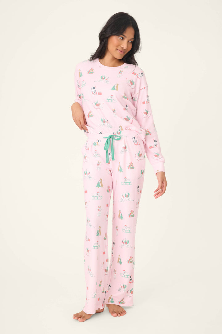 A woman stands barefoot, posing with one hand on her hip, wearing P.J. Salvage Peace & Pawlidays Dog Print Open Leg Pants and a matching pink long-sleeve top with playful animal prints. She looks off to the side.