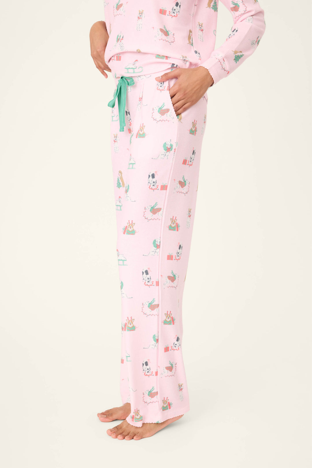 A person is shown from the waist down, barefoot against a plain background, wearing P.J. Salvage Peace & Pawlidays Dog Print Open Leg Pants with holiday-themed dog prints and a green drawstring.