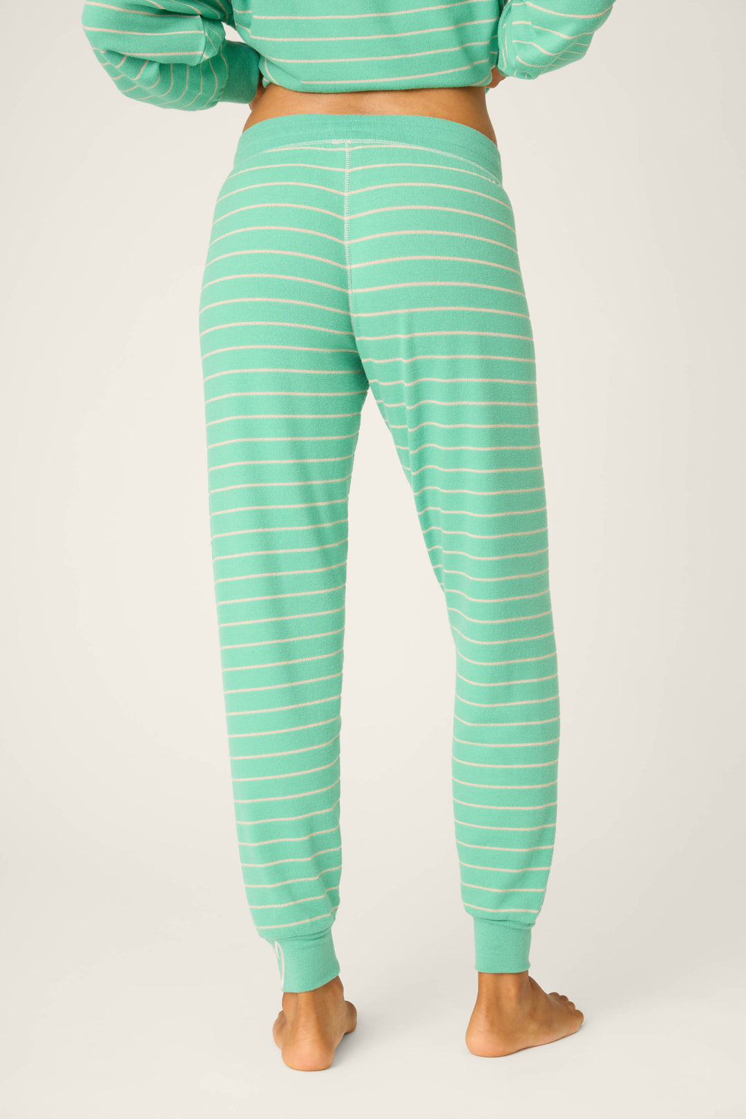 A person is shown from the back wearing P.J. Salvage Peace & Pawlidays Stripe Banded Pants—green joggers with white stripes, elastic waistband and cuffs—standing barefoot against a plain background.