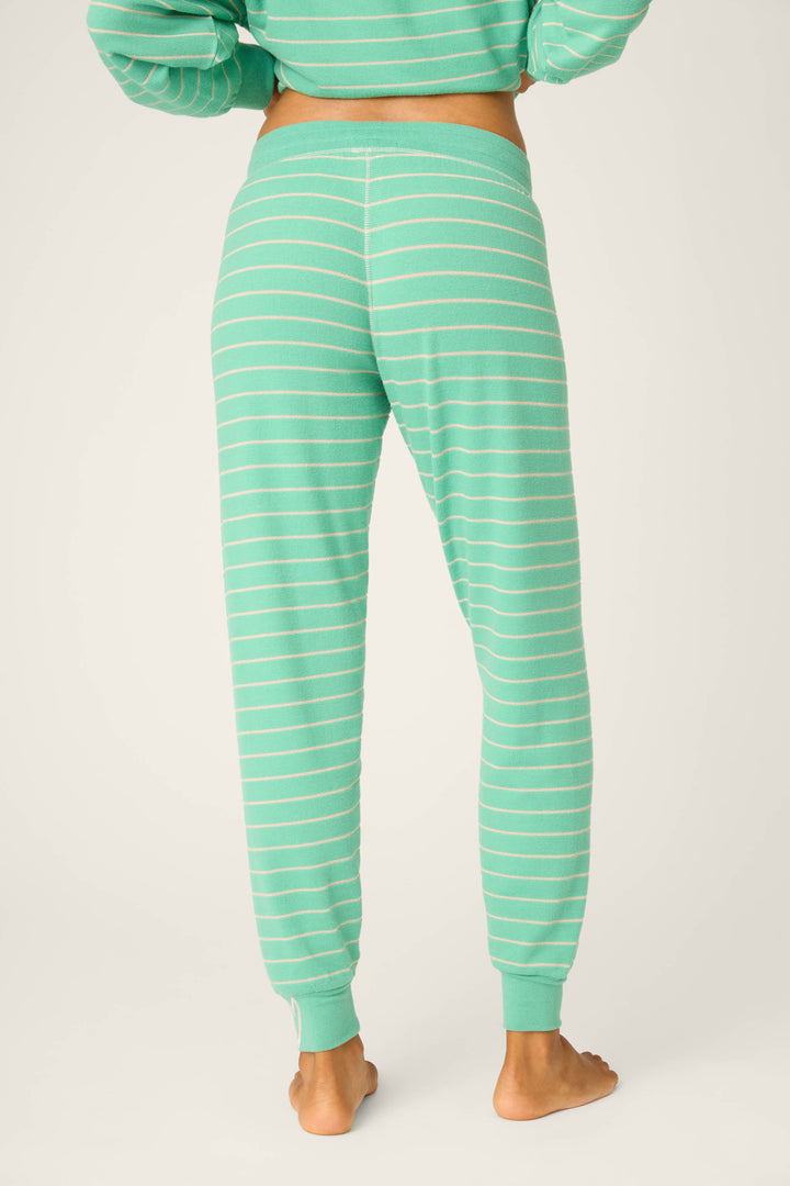 A person is shown from the back wearing P.J. Salvage Peace & Pawlidays Stripe Banded Pants—green joggers with white stripes, elastic waistband and cuffs—standing barefoot against a plain background.