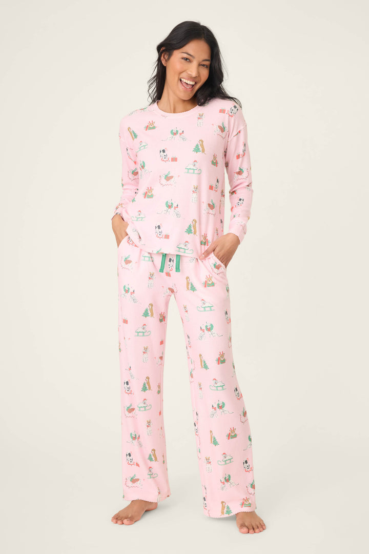 A woman stands barefoot against a plain background, smiling and wearing the P.J. Salvage Peace & Pawlidays Dog Print Pants Set, featuring playful holiday dog prints. Her hands are tucked into her pajama pants pockets.