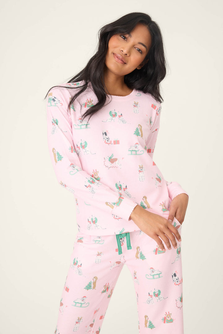 A woman with long dark hair wears the P.J. Salvage Peace & Pawlidays Dog Print Pants Set, a matching pink pajama set decorated with festive trees, sleds, and dogs, standing against a plain light background.