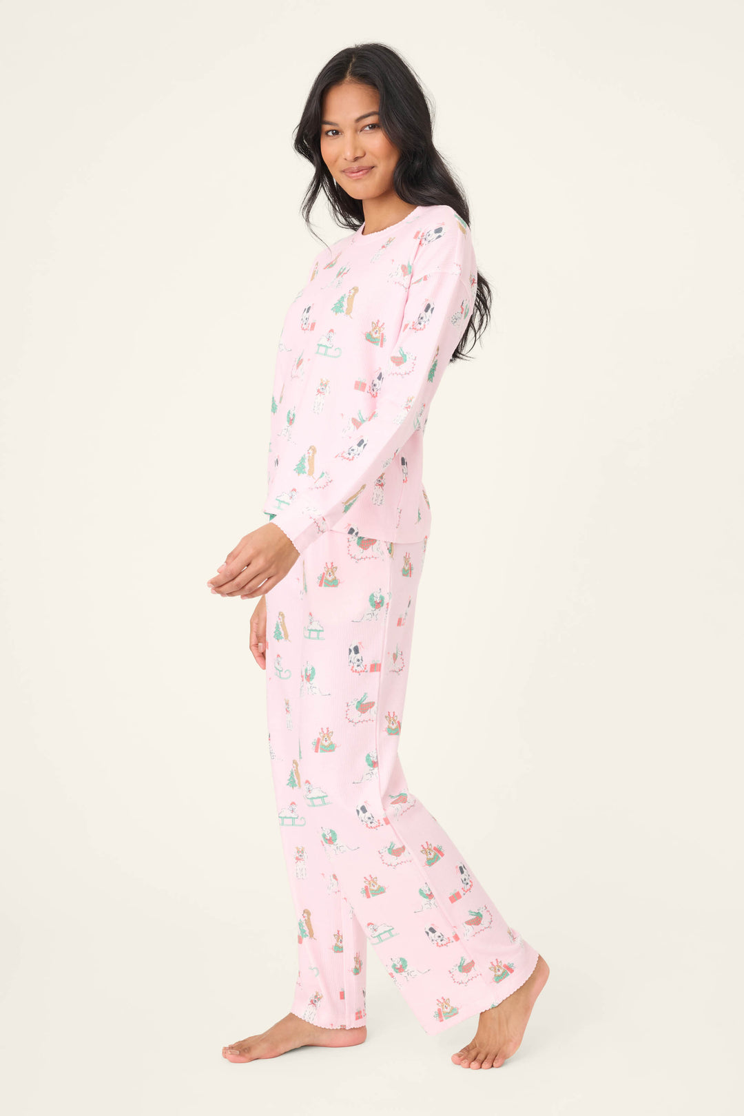 A person with long dark hair wears the Peace & Pawlidays Dog Print Pants Set by P.J. Salvage—light pink pajamas with a playful dog print—standing barefoot on a plain, off-white background and smiling at the camera.