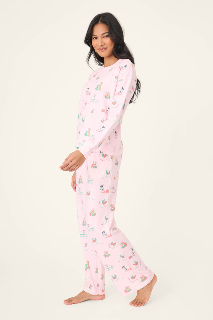 A person with long dark hair wears the Peace & Pawlidays Dog Print Pants Set by P.J. Salvage—light pink pajamas with a playful dog print—standing barefoot on a plain, off-white background and smiling at the camera.