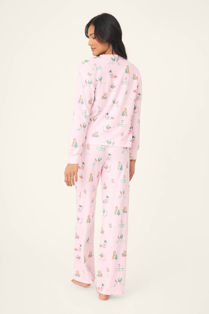 A woman stands barefoot facing away, wearing the P.J. Salvage Peace & Pawlidays Dog Print Pants Set—light pink, long-sleeve pajamas with a colorful dog print and full-length pants. The background is plain and light-colored.