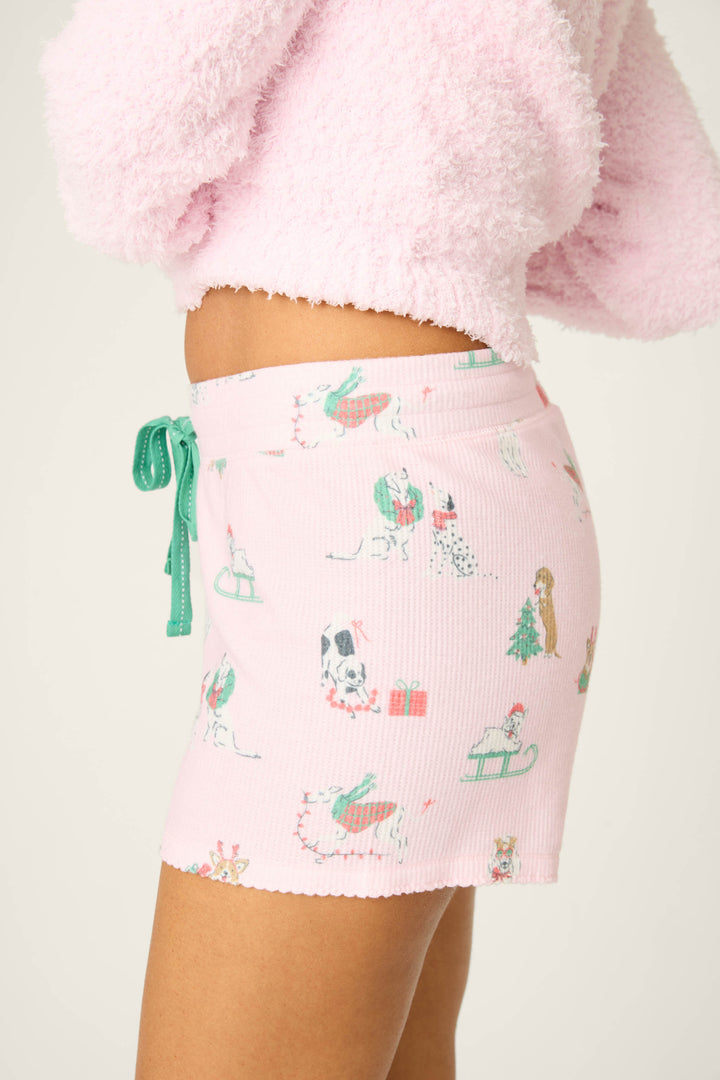 A person wears the P.J. Salvage DOGS SHORT PEACE & PAWLIDAYS pink pajama shorts with green drawstrings and festive dog designs, paired with a fuzzy light pink long-sleeve top, photographed from the side against a plain backdrop.