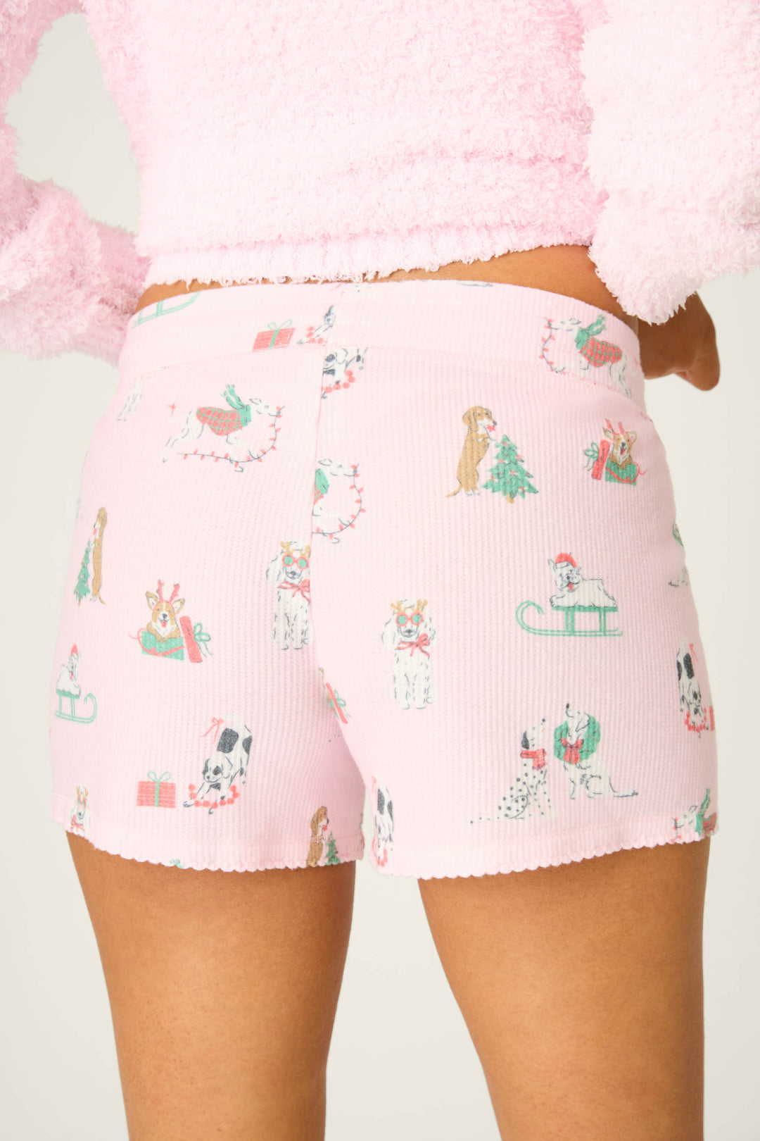 A person is wearing P.J. Salvage DOGS SHORT PEACE & PAWLIDAYS—light pink shorts with cartoon dogs, Christmas trees, presents, and sleds. Their fluffy pink top is cropped out except for the sleeves and lower hem.