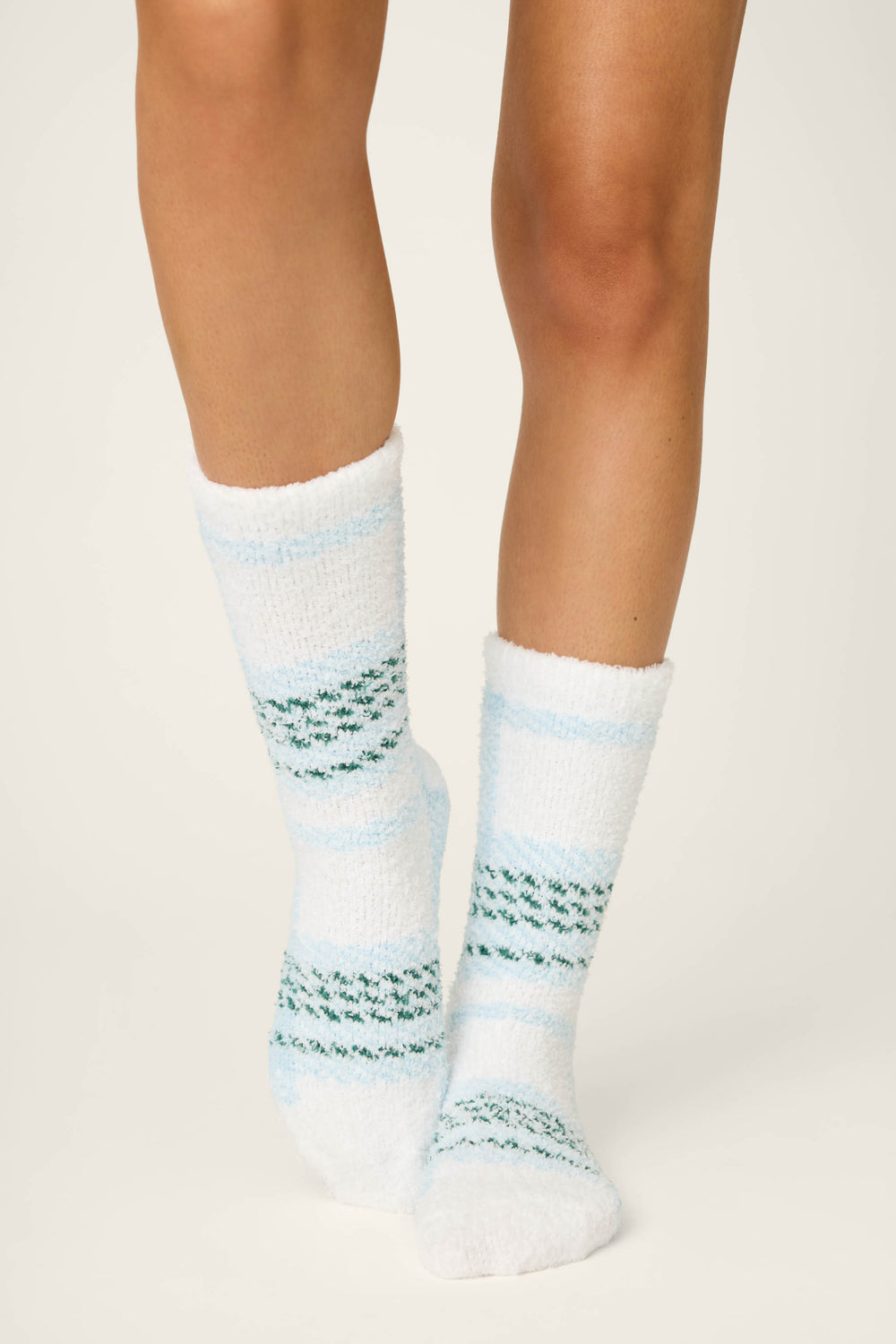 Close-up of a person's lower legs wearing P.J. Salvage PLAID SOCKS WINTER WONDERLAND—fuzzy white socks with light blue and green stripes—standing on a plain, light-colored surface.