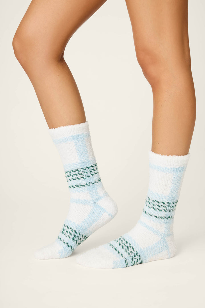 A person wears P.J. Salvage PLAID SOCKS WINTER WONDERLAND in cozy white and light blue plaid with green accents; only their lower legs and feet show against a plain light background.