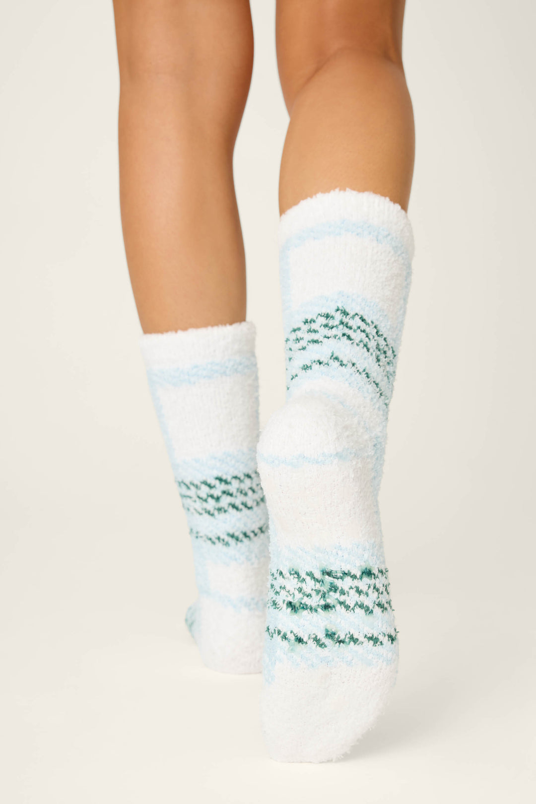 A person stands on tiptoe, displaying the backs of their lower legs and feet in P.J. Salvage PLAID SOCKS WINTER WONDERLAND—white, fuzzy socks with light blue and green stripes—against a plain light background.