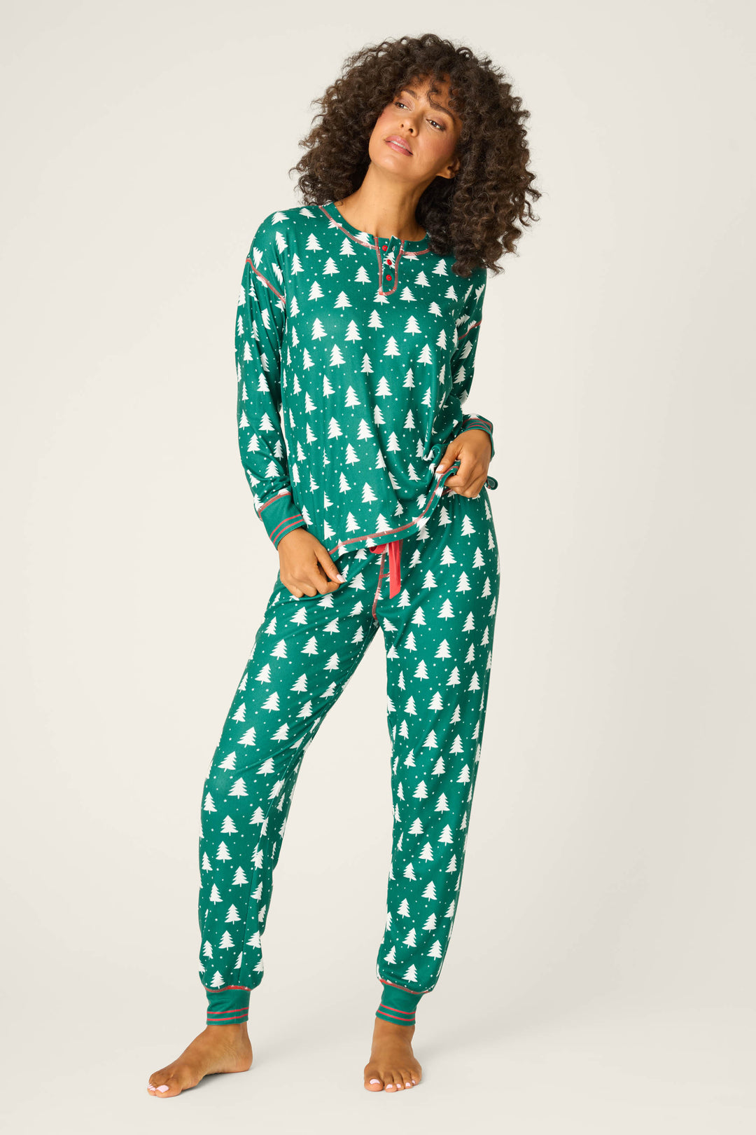 A person models the P.J. Salvage 3-Piece Tree Print Pointelle PJ Set—forest green with white tree prints and red accents—standing barefoot with a hand on their hip, showing off perfect holiday pajamas for cozy nights.