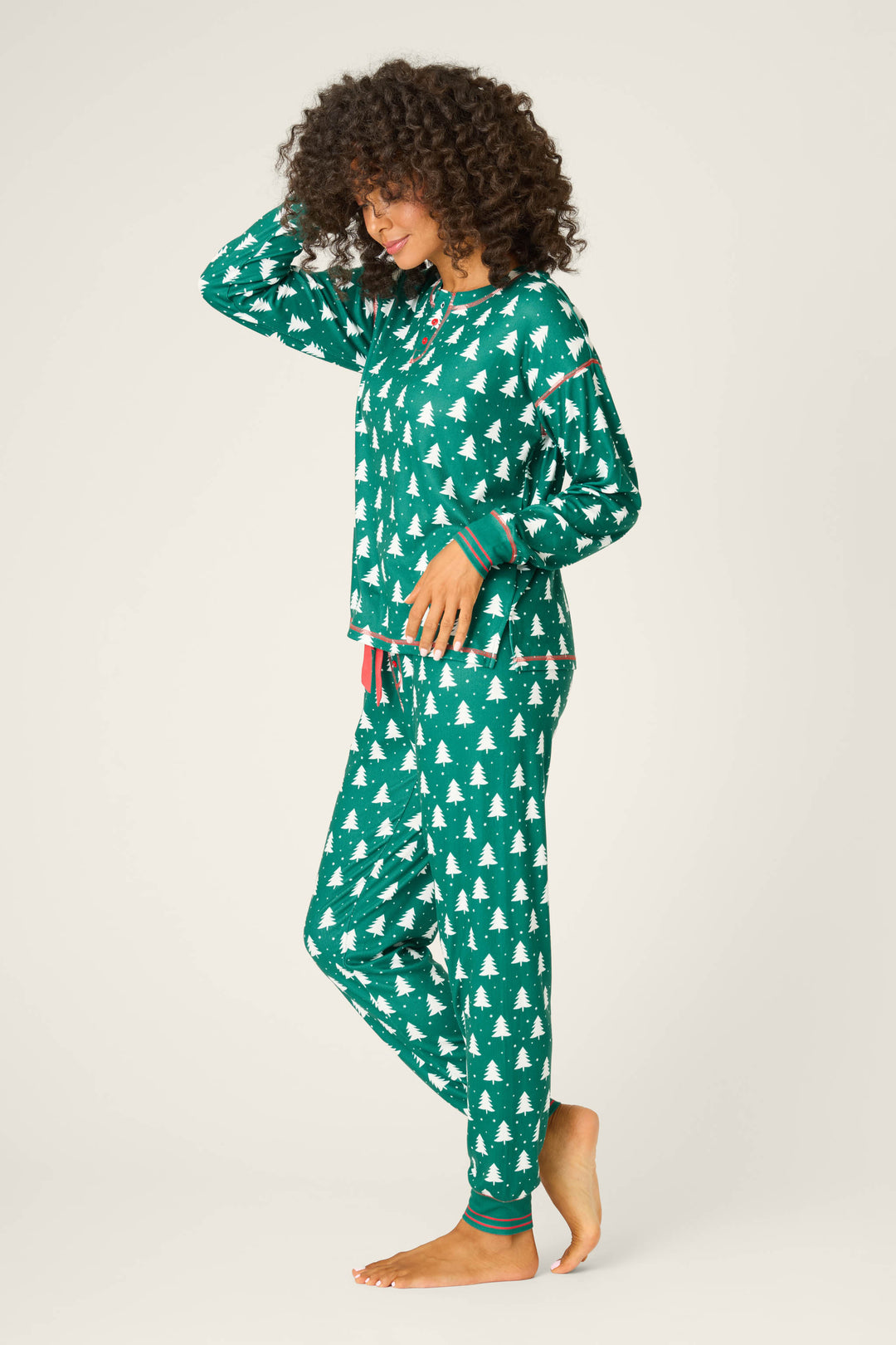 A person stands barefoot on a plain background, smiling in the P.J. Salvage 3-Piece Tree Print Pointelle PJ Set, featuring a forest-green color with white Christmas tree motifs. Their curly hair frames their face as they pose with one hand on their head.