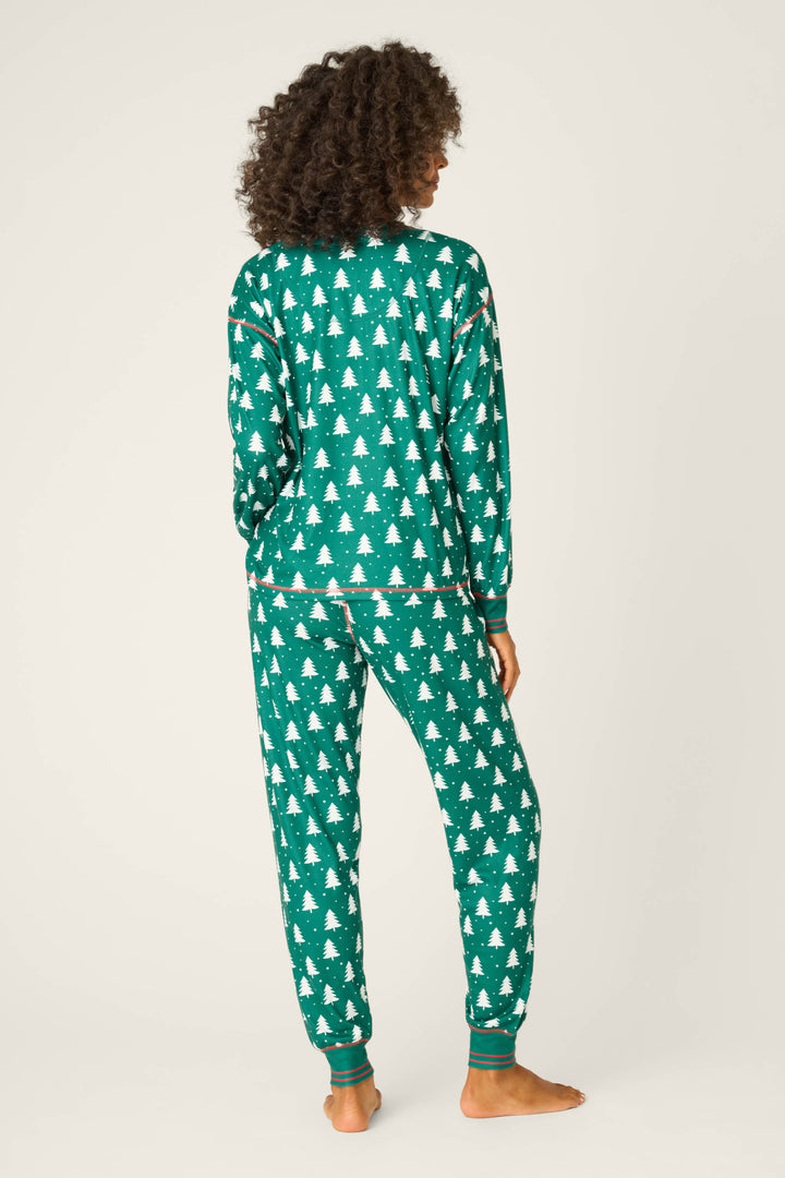 A person stands barefoot facing away, wearing the P.J. Salvage 3-Piece Tree Print Pointelle PJ Set featuring a forest-green top and pants with a white Christmas tree pattern, perfect holiday pajamas against an off-white background.
