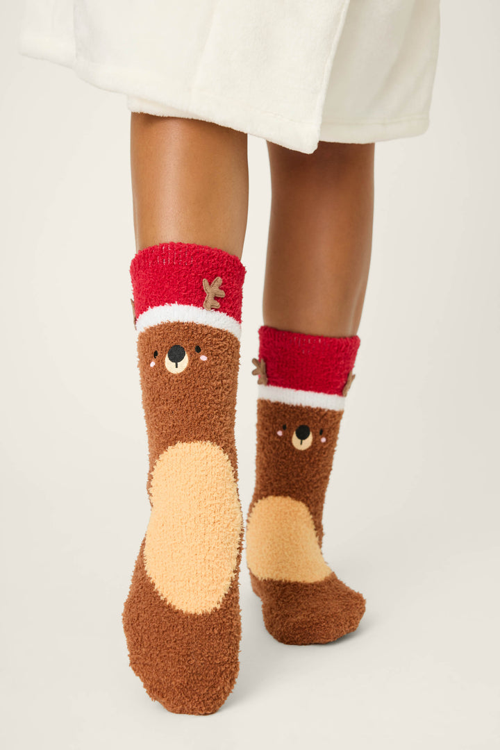 A person in a white robe stands on a light surface, wearing P.J. Salvage REINDEER SOCKS SKI JAMMIE CLASSICS—fuzzy brown socks with red cuffs, featuring a reindeer face and antlers design on the front.