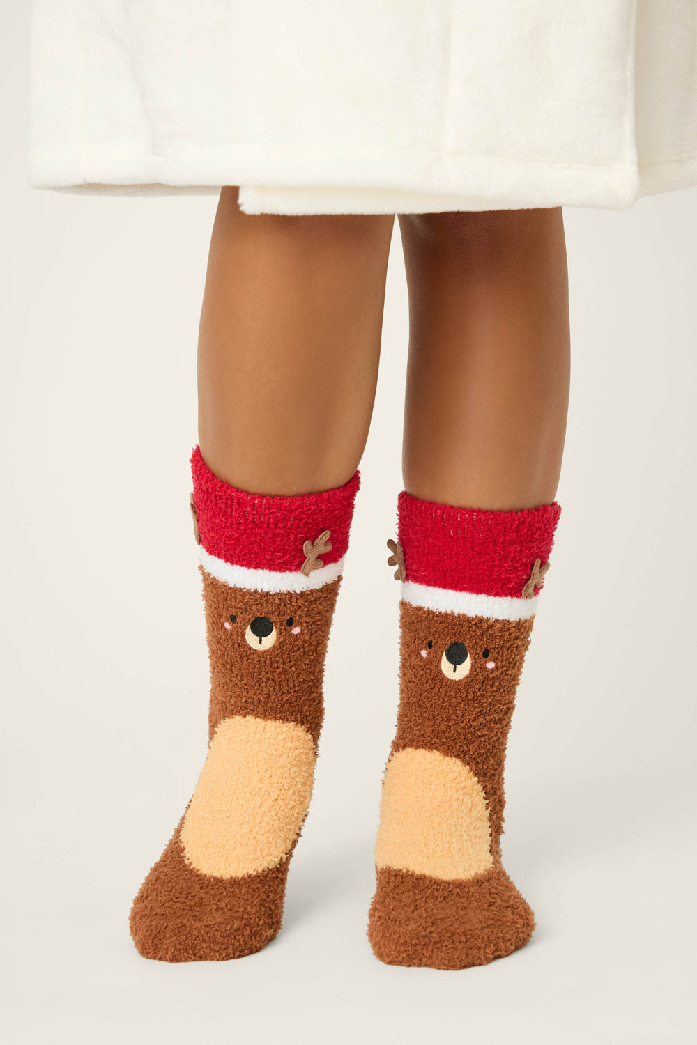 A person in a white robe wears P.J. Salvage REINDEER SOCKS SKI JAMMIE CLASSICS—fuzzy brown socks with red cuffs, each featuring a reindeer face and antlers.