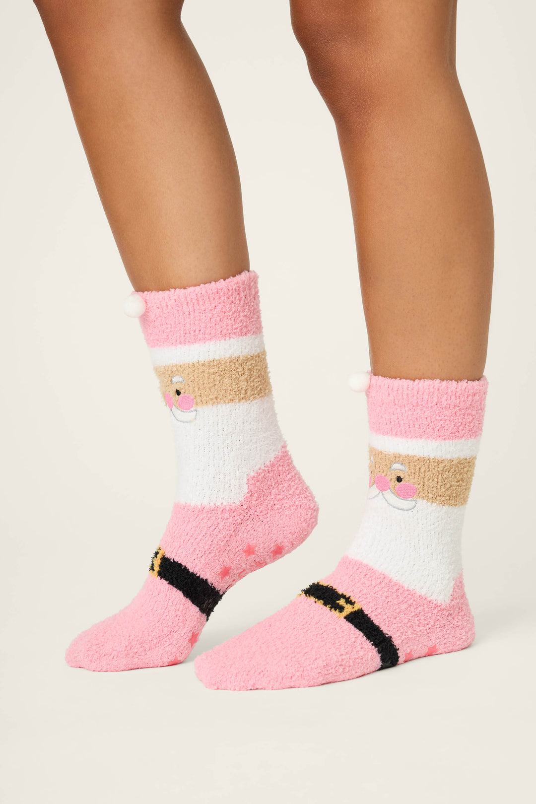 A person wears SANTA SOCKS SKI JAMMIE CLASSICS by P.J. Salvage—fluffy pink and white socks with a cartoon Santa face and black belt detail—against a plain background.