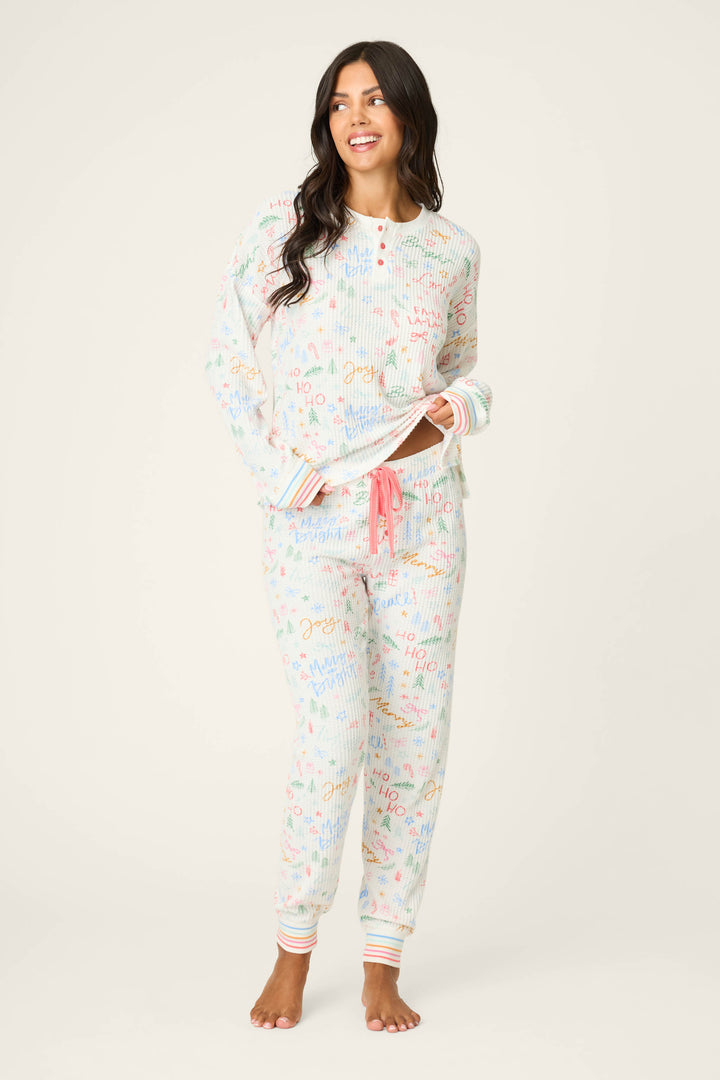 A woman stands barefoot in the P.J. Salvage Ski Jammie Holiday Print PJ Set, a matching white pajama set featuring colorful science-themed patterns, a button-up long-sleeve top, and jogger pants with a pink drawstring and striped cuffs.