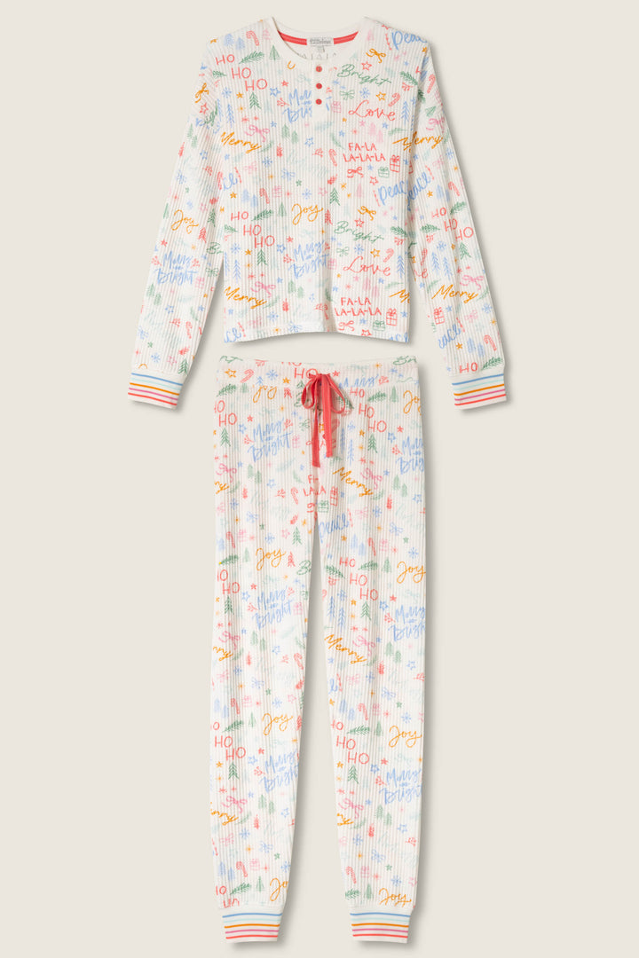 The P.J. Salvage Ski Jammie Holiday Print PJ Set features a white button-up long-sleeve top and drawstring pants adorned with colorful holiday text and doodles, plus striped cuffs on both the sleeves and pant legs.
