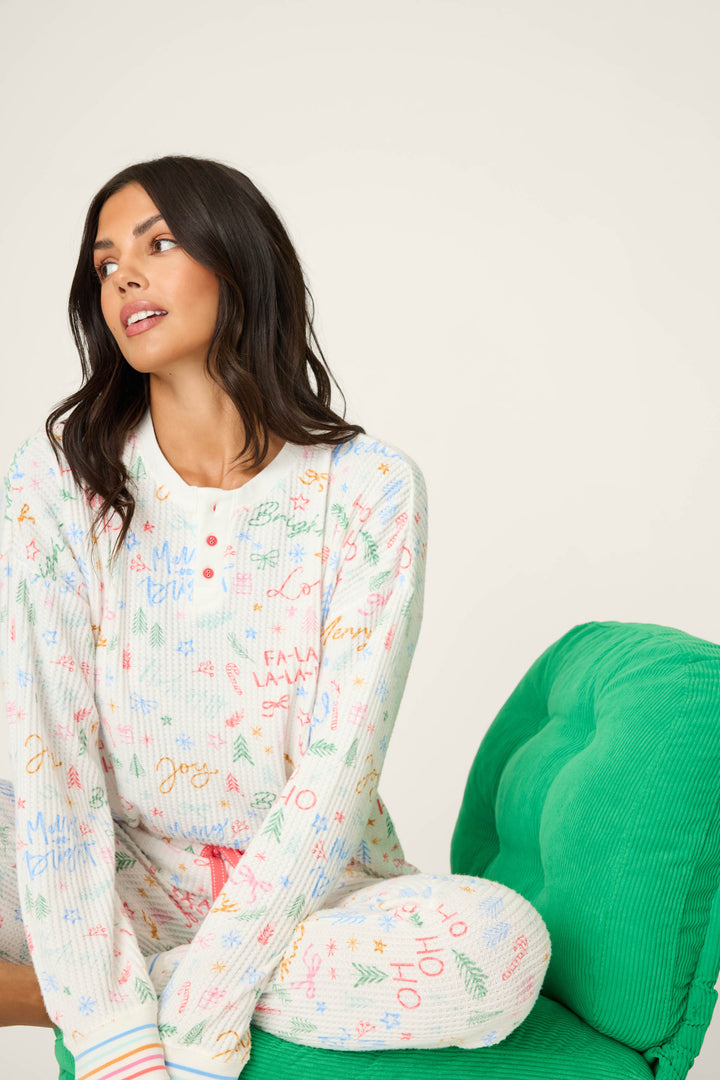 A woman with long dark hair sits sideways on a bright green chair, wearing the P.J. Salvage Ski Jammie Holiday Print PJ Set, featuring festive multicolored words and symbols. She looks to her left with a relaxed expression.