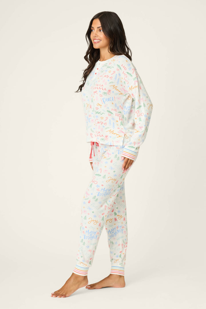 A woman stands barefoot on a plain background, wearing the P.J. Salvage Ski Jammie Holiday Print PJ Set with colorful cursive writing and striped cuffs, facing to the side and smiling slightly.