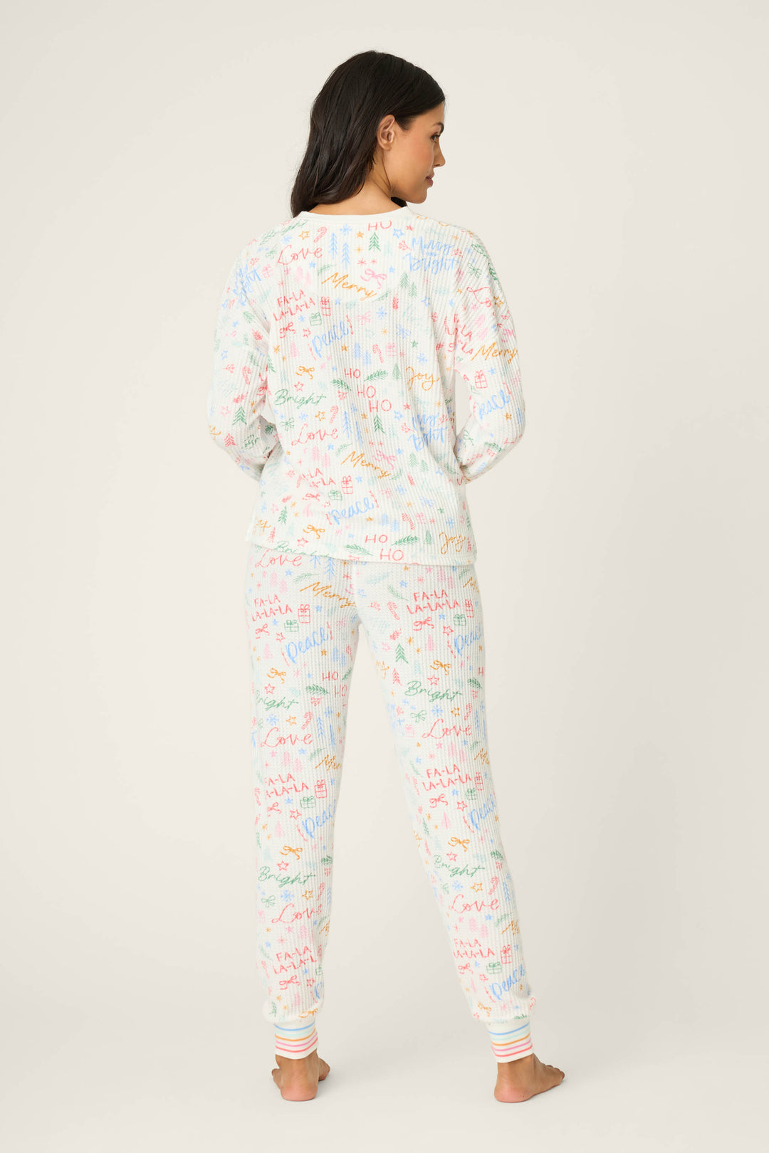 A woman stands barefoot facing away, wearing the P.J. Salvage Ski Jammie Holiday Print PJ Set—white long-sleeve pajamas and pants with colorful doodle patterns—against a plain light background.