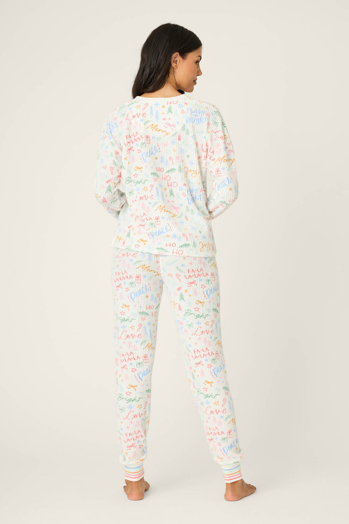 A woman stands barefoot facing away, wearing the P.J. Salvage Ski Jammie Holiday Print PJ Set—white long-sleeve pajamas and pants with colorful doodle patterns—against a plain light background.
