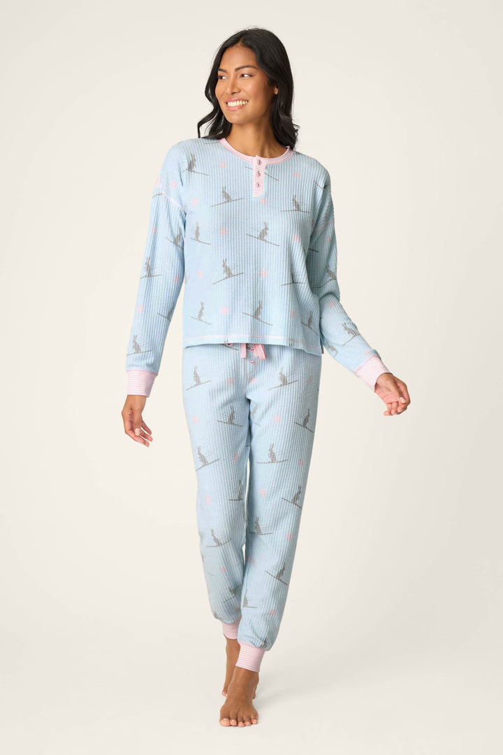 A woman stands barefoot, smiling and looking to the side in the P.J. Salvage Ski Jammie Classics Ski Bunny PJ Set—light blue, long-sleeved pajamas with pink-trimmed collar and cuffs, subtle printed design, and ribbed texture.