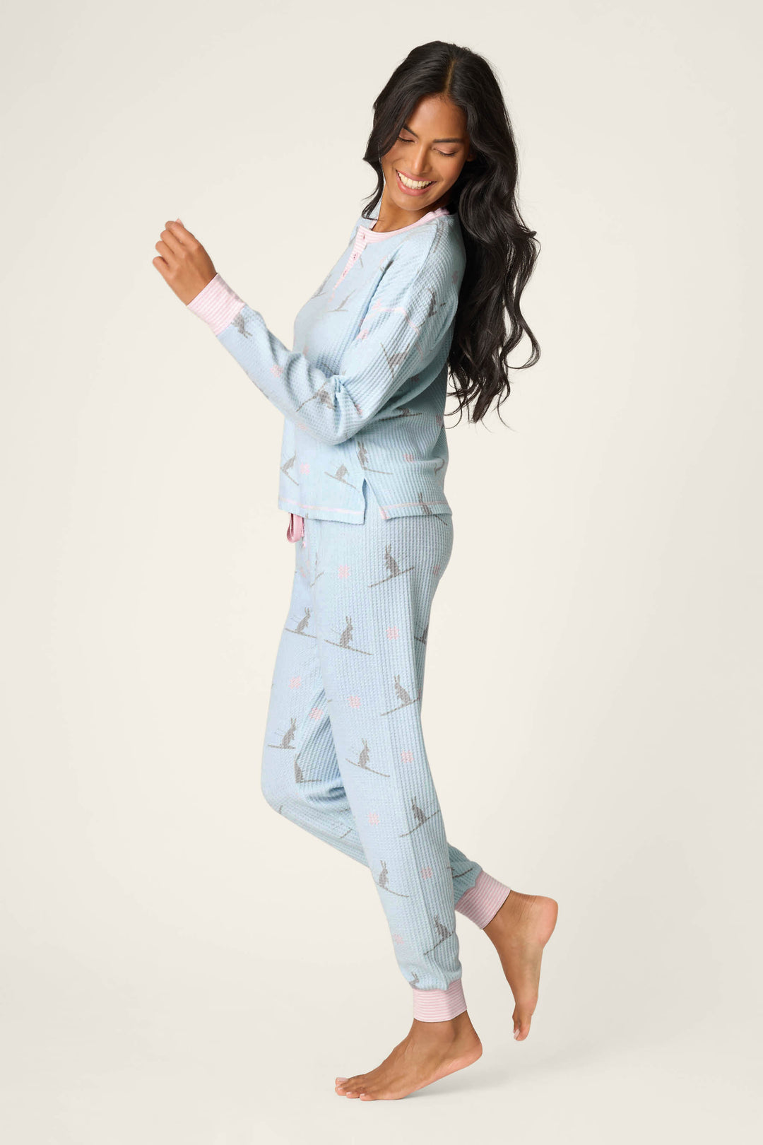 A woman with long dark hair smiles while standing on one foot, wearing the P.J. Salvage Ski Jammie Classics Ski Bunny PJ Set—light blue pajamas with a subtle print and pink cuffs—against a plain light background.