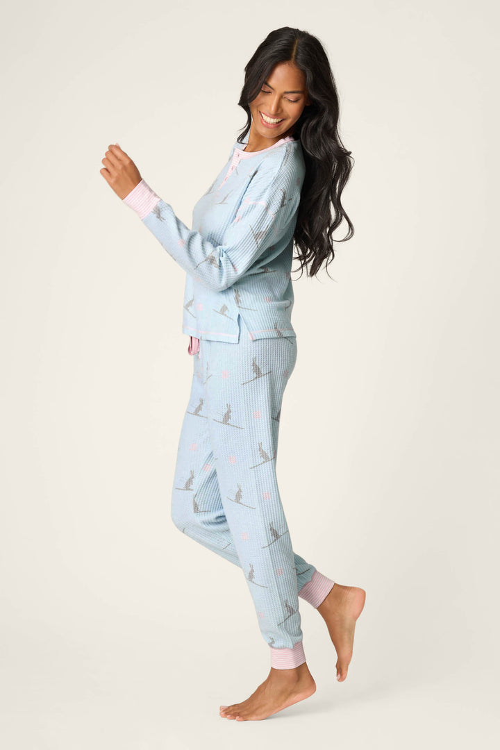 A woman with long dark hair smiles while standing on one foot, wearing the P.J. Salvage Ski Jammie Classics Ski Bunny PJ Set—light blue pajamas with a subtle print and pink cuffs—against a plain light background.