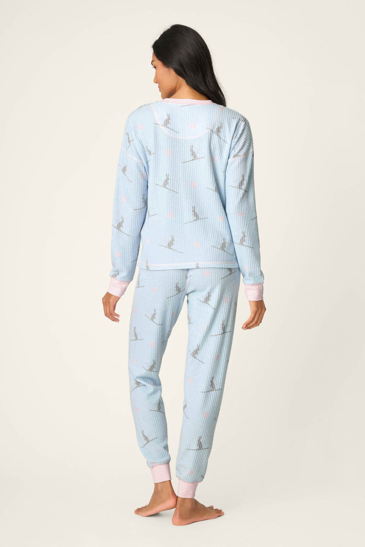 A person with long dark hair stands barefoot, facing away while wearing the P.J. Salvage Ski Jammie Classics Ski Bunny PJ Set—light blue ribbed pajamas featuring small gray figures, pink details, and light pink cuffs at the wrists and ankles.