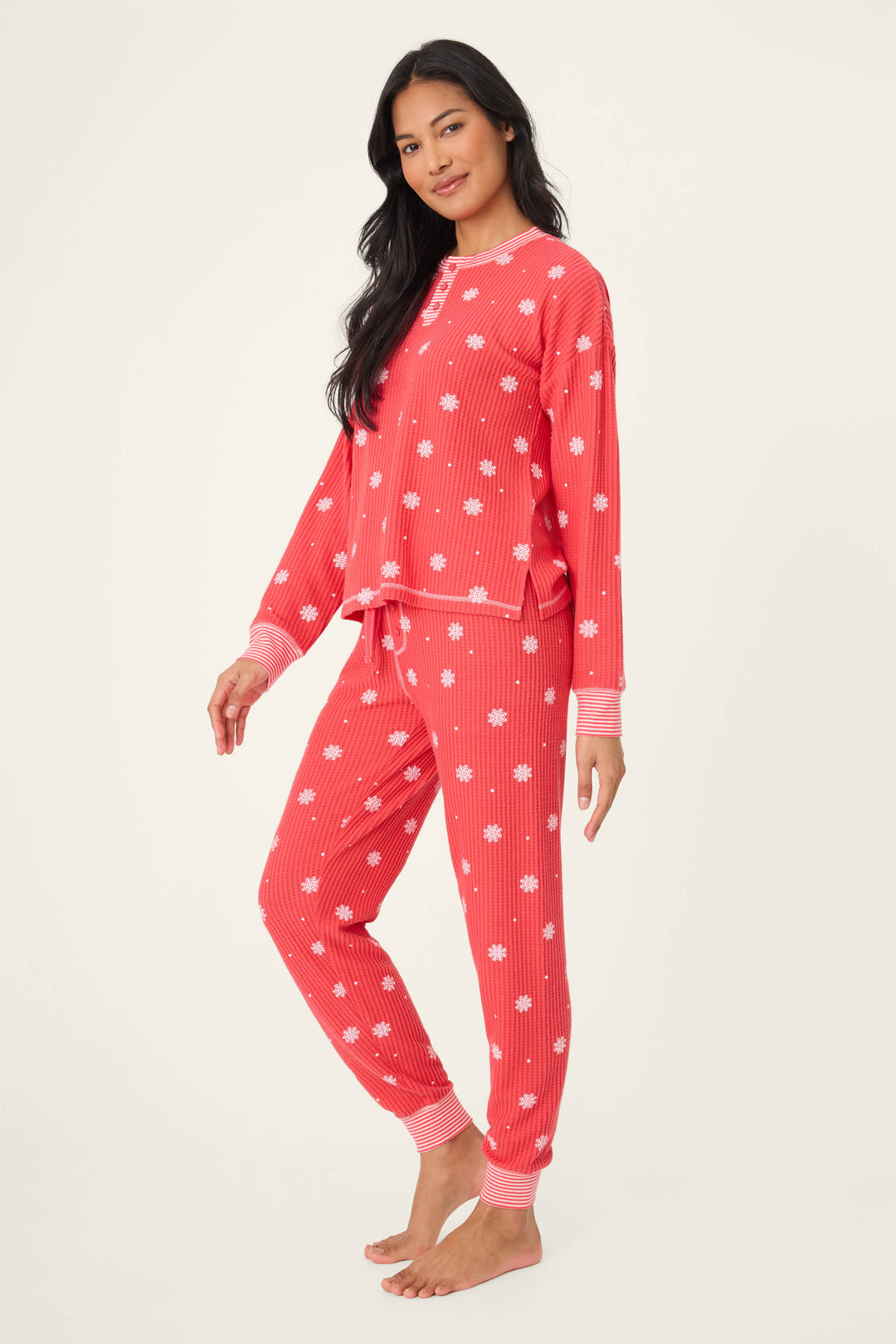 A woman wears the P.J. Salvage Ski Jammie Snowflake Print PJ Set—barefoot, in the matching button-up long-sleeve top and elastic-waist pants, smiling slightly at the camera.