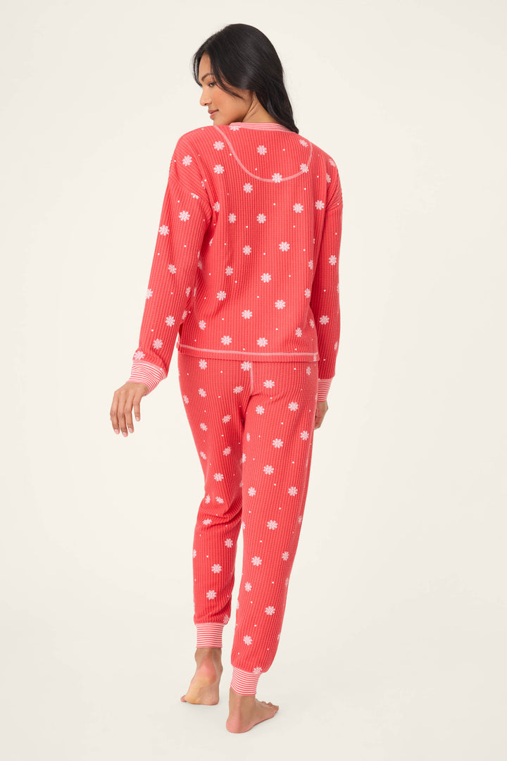 A person stands barefoot facing away, wearing the P.J. Salvage Ski Jammie Snowflake Print PJ Set, featuring a red base with white snowflake prints and pink cuffs, against a plain light background.