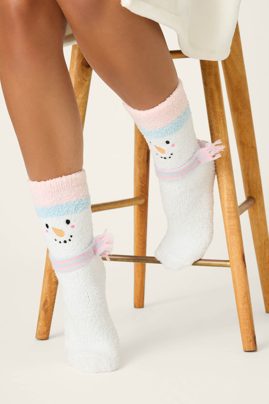 A person sits on a wooden stool wearing P.J. Salvage SNOWMAN SOCKS SKI JAMMIE CLASSICS—fuzzy white socks with pastel pink and blue bands and a snowman face near the ankle. Only their lower legs and feet are visible.