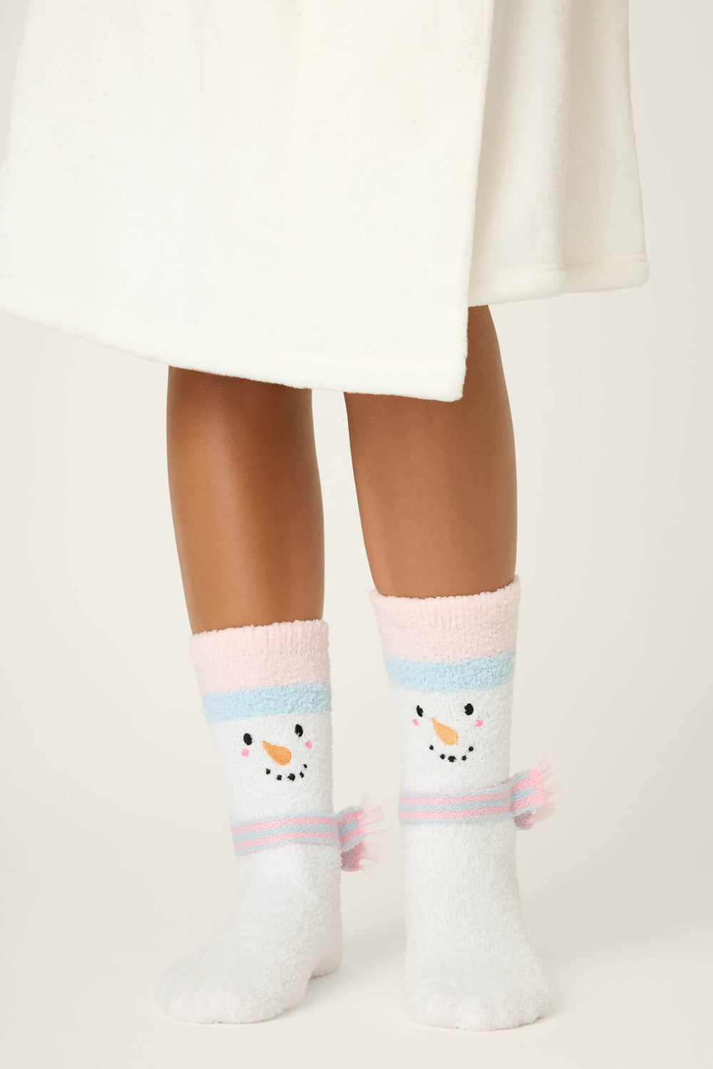 A person in a white robe stands on a light surface, wearing P.J. Salvage SNOWMAN SOCKS SKI JAMMIE CLASSICS featuring fuzzy white fabric, pastel pink and blue stripes, and snowman faces on the front.