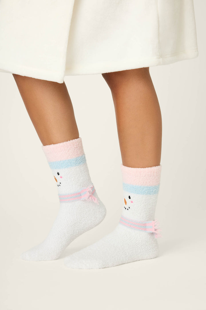 A person wearing P.J. Salvage SNOWMAN SOCKS SKI JAMMIE CLASSICS—white fuzzy socks with pastel pink and blue stripes, a small animal face, and pink bows—stands on a light background with part of a white garment visible.