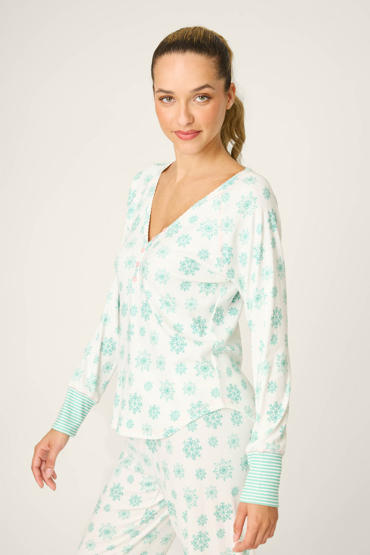 A person faces forward against a light background, wearing the P.J. Salvage Snowflake Wishes Pointelle Henley Top—white with blue snowflakes, blue striped cuffs, V-neck, and long sleeves; hair is tied back.