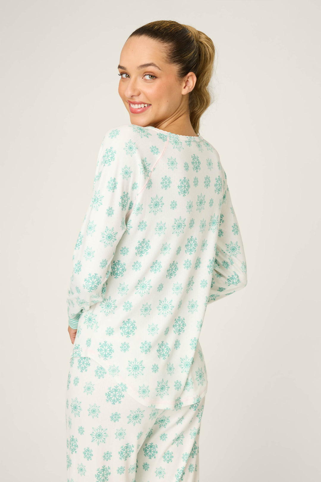A woman with light brown hair in a ponytail smiles at the camera, wearing the P.J. Salvage Snowflake Wishes Pointelle Henley Top—white sleepwear with a light blue snowflake pattern—against a plain background.