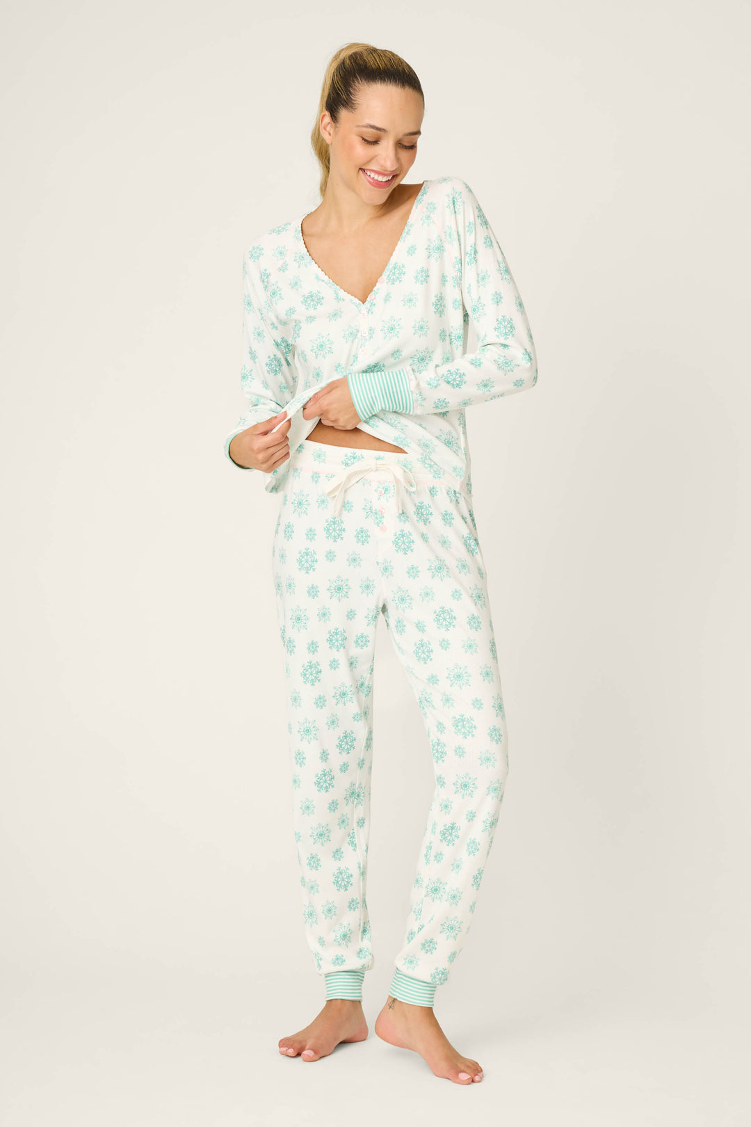 A woman stands barefoot on a light background, smiling as she lifts her pajama top, wearing P.J. Salvage Snowflake Wishes Pointelle Banded Pants with a blue snowflake pattern.