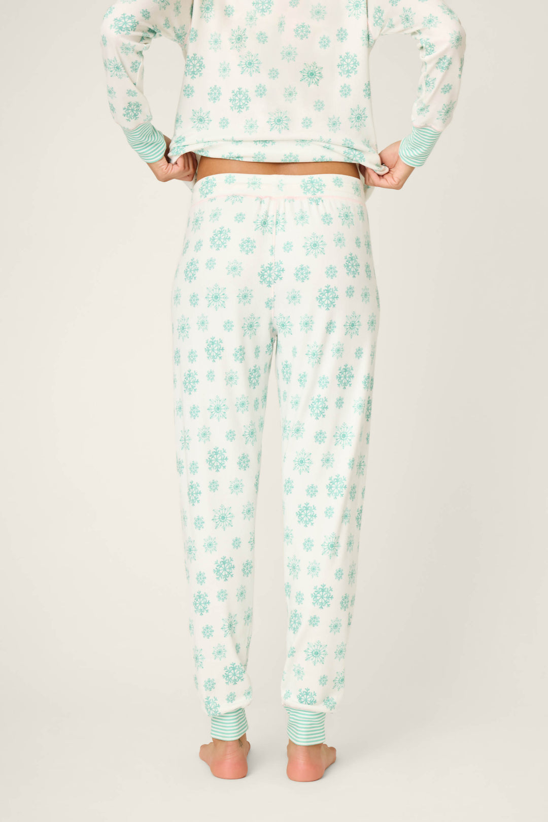 A person stands barefoot, facing away, wearing P.J. Salvage Snowflake Wishes Pointelle Banded Pants in white with a light blue snowflake pattern and striped cuffs, paired with a matching long-sleeve top against a plain light background.