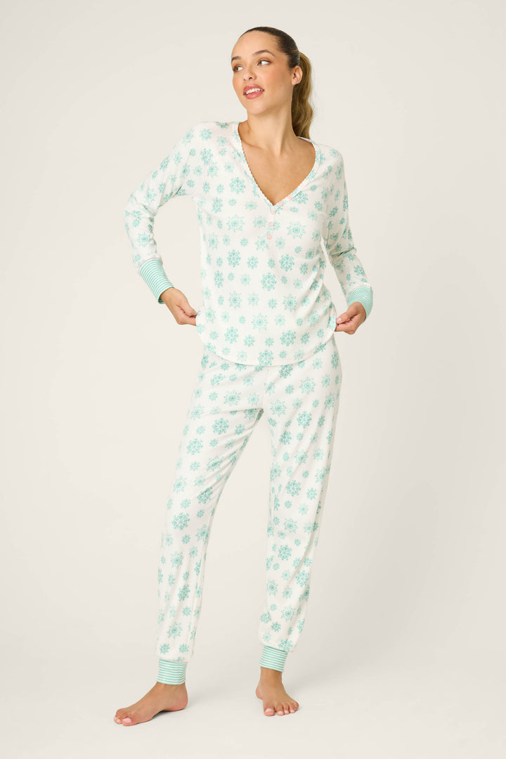 A woman stands barefoot against a plain background, wearing the P.J. Salvage Snowflake Wishes Pointelle Pants Set—a light blue pajama with snowflake pattern, long sleeves and pants with blue cuffs. She has her hands in her pockets and looks to the side.