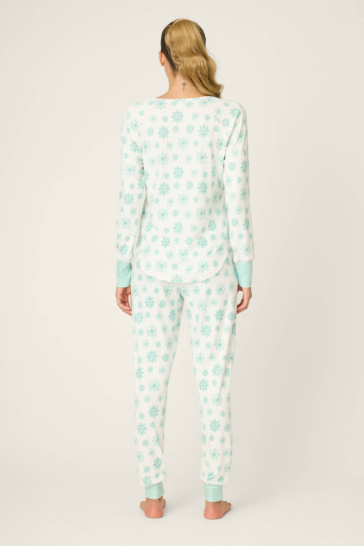 A person stands barefoot facing away, wearing the P.J. Salvage Snowflake Wishes Pointelle Pants Set—white brushed pointelle fabric with light blue snowflake patterns and matching light blue striped cuffs on the sleeves and ankles.