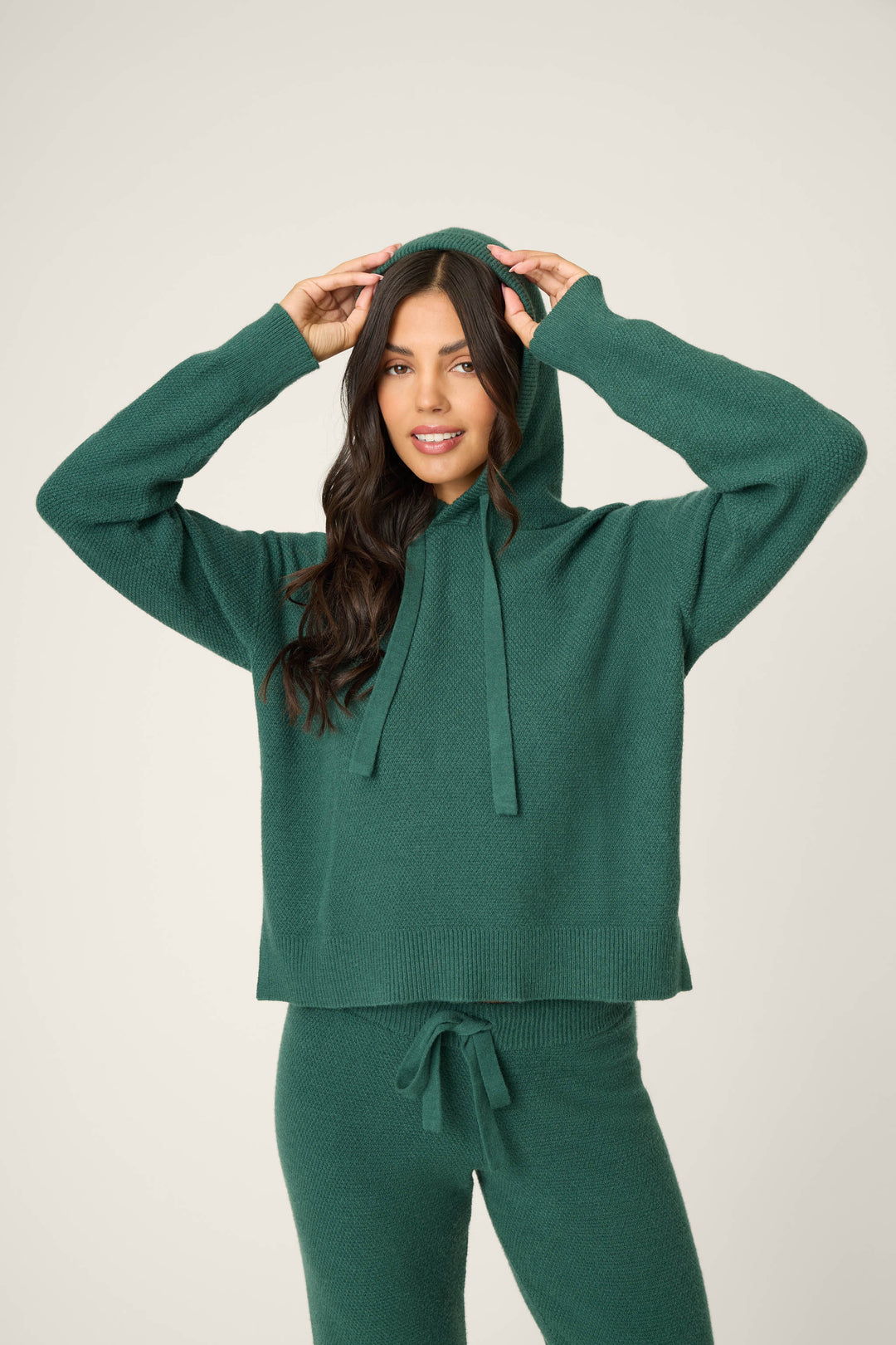 A woman with long dark hair smiles slightly while facing forward in a P.J. Salvage SOLID PJ SET SWEATER WEATHER—dark green knit hoodie and matching pants. She holds the hood with both hands against a plain, light background.